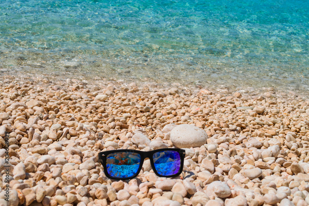 Blue sunglasses on a pebble beach with turquoise sea waters on the background. Summer holiday concept 