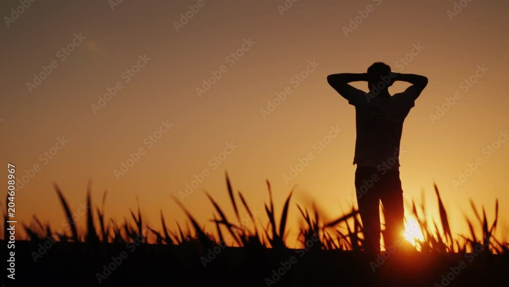 Silhouette A young man enjoys the fresh air and admires the sunset. He holds his hands behind the back of his head, looks ahead in front of him