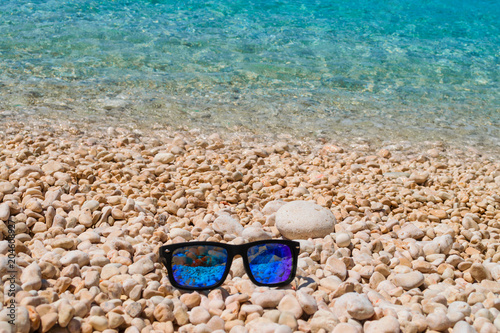 Blue sunglasses on a pebble beach with turquoise sea waters on the background. Summer holiday concept 