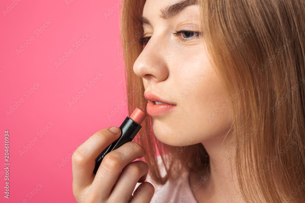 A young girl paints lips with lipstick and looking in the mirror , on a pink background, isolated, fun and beautiful A young girl paints lips with lipstick and looking in the mirror , on a pink background, isolated, fun and beautiful