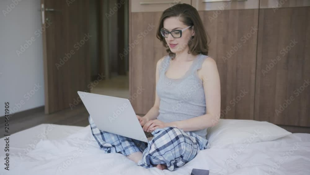 Beautiful young woman in pajamas browsing laptop while sitting on comfortable mattress in stylish bedroom in morning. Beautiful young woman in pajamas browsing laptop while sitting on comfortable mattress in stylish bedroom in morning.