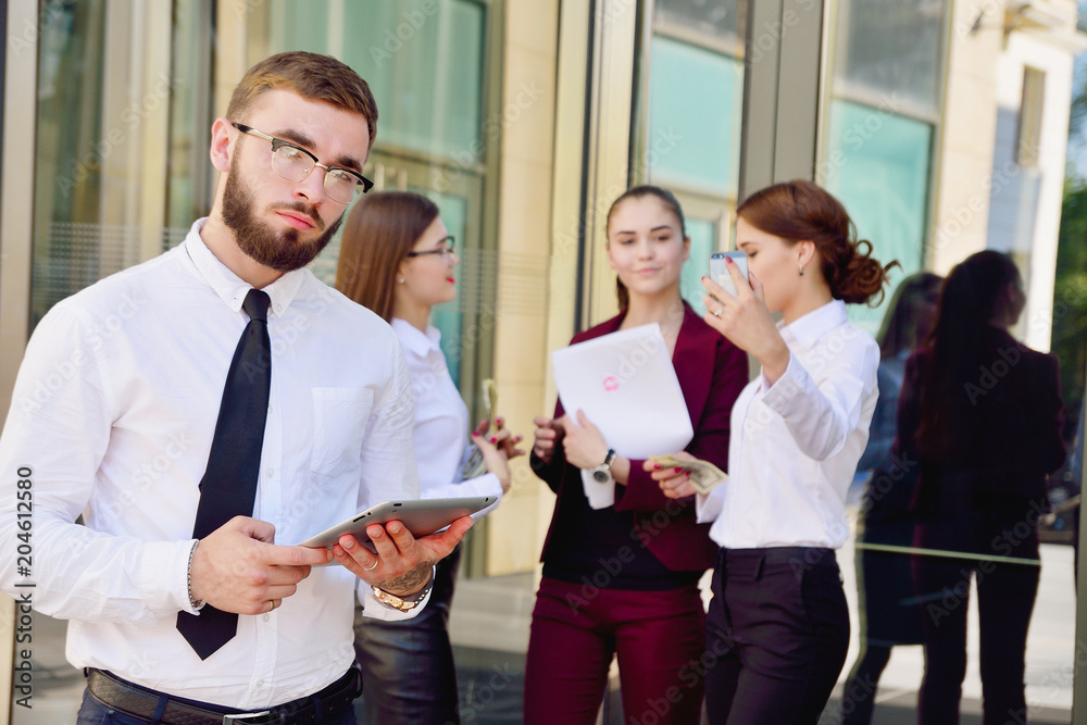 A young male manager with a tablet in his hands against the background of the girls. Office staff in the background of a multi-storey glass building. Lifestyle. Businessman A young male manager with a tablet in his hands against the background of the girls. Office staff in the background of a multi-storey glass building. Lifestyle. Businessman