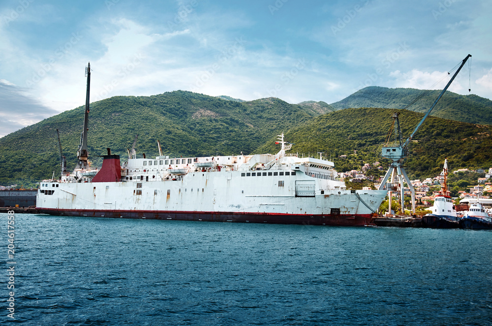 old ship went to the Adriatic Shipyard Bijela for maintenance and refit. .Landscape of ships, tugboats and cranes in shipyard in Boka Kotorska Bay opposite small town in mountains.