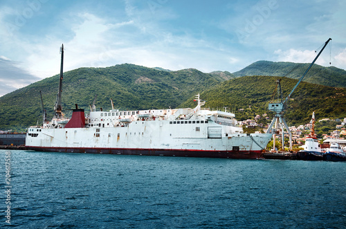 old ship went to the Adriatic Shipyard Bijela for maintenance and refit. .Landscape of ships, tugboats and cranes in shipyard in Boka Kotorska Bay opposite small town in mountains.
