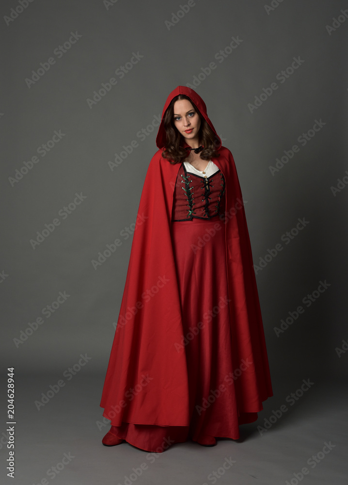 full length portrait of woman wearing red fantasy costume with cloak, standing pose on grey studio background.