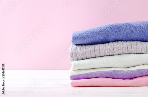 Stack of colorful perfectly folded sweaters on white wood texture table. Pile of different pastel color shirts and sweaters on wooden table, pale pink wall background. Close up, top view, copy space.