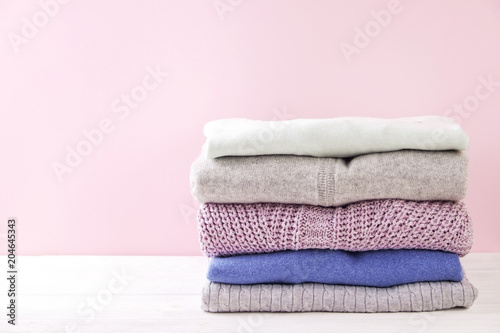 Stack of colorful perfectly folded sweaters on white wood texture table. Pile of different pastel color shirts and sweaters on wooden table, pale pink wall background. Close up, top view, copy space.