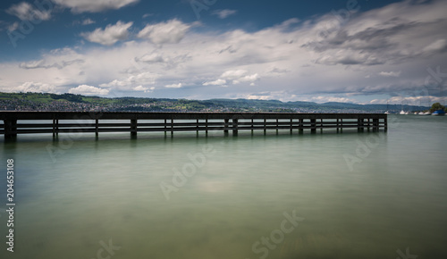 wooden pier on lake Zurich with rolling hills mountain landscape and sailboats in the background