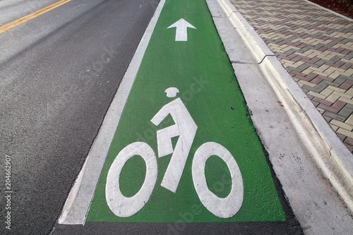Green Bicycle Lane with Arrow Pointing Up Ahead Alongside Road with Double Yellow Lines to the Left and Tiled Sidewalk to the Right Back-Lit by the Sun