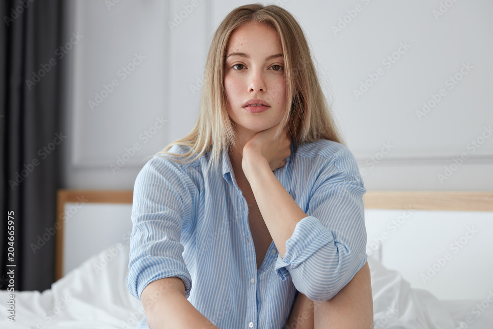 Naklejka premium Pretty young female with pensive look sits on bed in modern hotel room, wears striped shirt, plans her travel, feels refreshed after healthy sleep, feels comfort, enjoys good service. Bed time concept