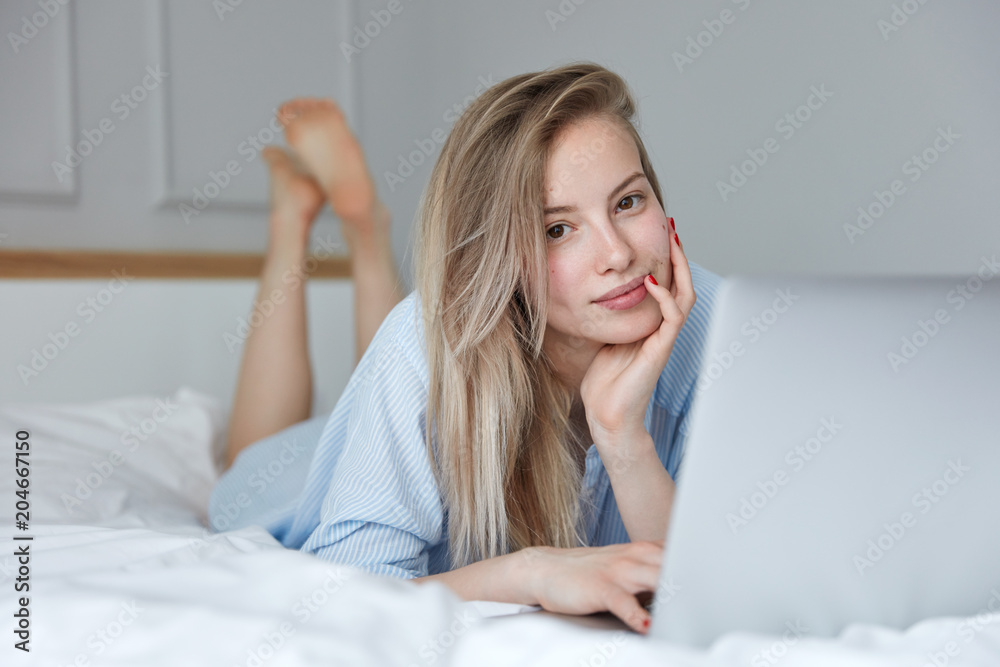 Adorable female freelancer in casual striped night wear, lies on white bed in morning, works remotely on portable laptop computer, promots social media account. People, leisure and technology Adorable female freelancer in casual striped night wear, lies on white bed in morning, works remotely on portable laptop computer, promots social media account. People, leisure and technology