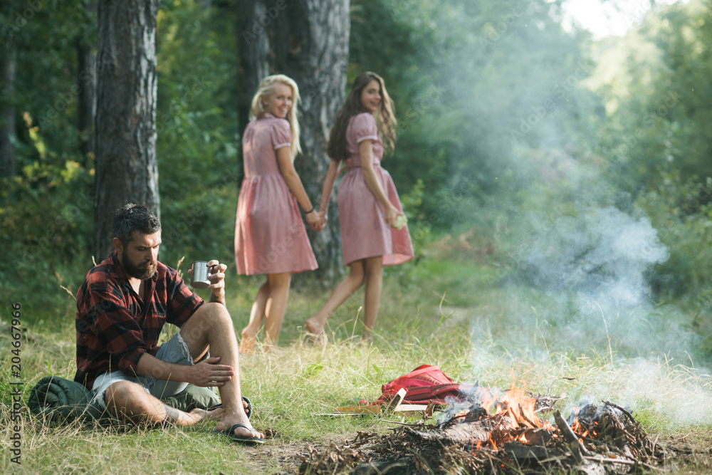 Two happy smiling girls looking back at thoughtful man sitting on grass. Bearded man in lumberjack shirt thinking while looking at flames of campfire. Nostalgia and memories concepts