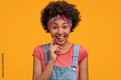 Cute happy hipster woman with positive expression, wears fashionable overalls and headband, being in good mood after party with friends, expresses positive emotions, isolated on yellow background