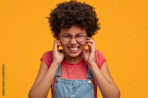 Portrait of annoyed frustrated American female plugs ears and clenches teeth, being irritated with annoying loud sound, isolated over yellow background. People, problems and emotions concept.