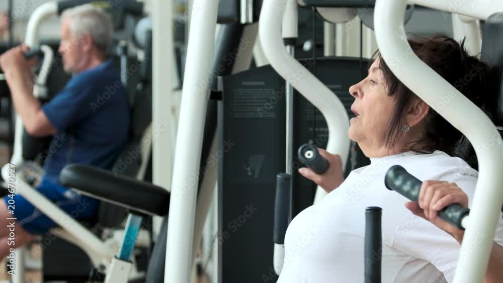 Female pensioner training at gym, side view. Attractive senior woman working out at fitness center close up. Sport, wellness and healthcare. Female pensioner training at gym, side view. Attractive senior woman working out at fitness center close up. Sport, wellness and healthcare.