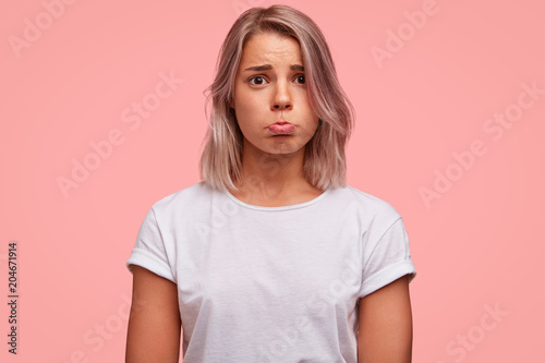 Fotografie, Tablou Abused pretty female curves lower lip, being offended during quarrel, looks with sorrowful miserable expression, wears casual white t shirt, isolated over pink background