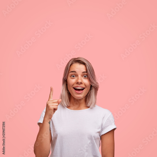 Beautiful female with excited look, wears brackets and white t shirt, indicates at blank copy space over pink background, sees something amazing. Joyful woman with bobbed hairstyle gestures.