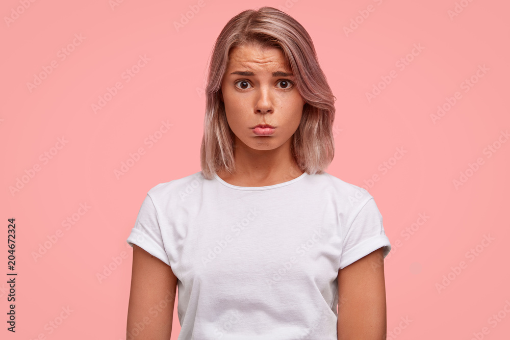 Waist up portrait of unahappy upset femal model in white casual clothes, curves lower lip with dissatisfaction, has sullen expression, isolated over pink background. Negative human emotions concept Waist up portrait of unahappy upset femal model in white casual clothes, curves lower lip with dissatisfaction, has sullen expression, isolated over pink background. Negative human emotions concept
