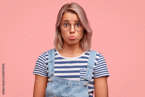 Cute adorable woman with unhappy expression, curves lips as being victim of sexual abusement, wears casual clothes, isolated over pink studio background. Discontent Caucasian girl with dyed hair photo