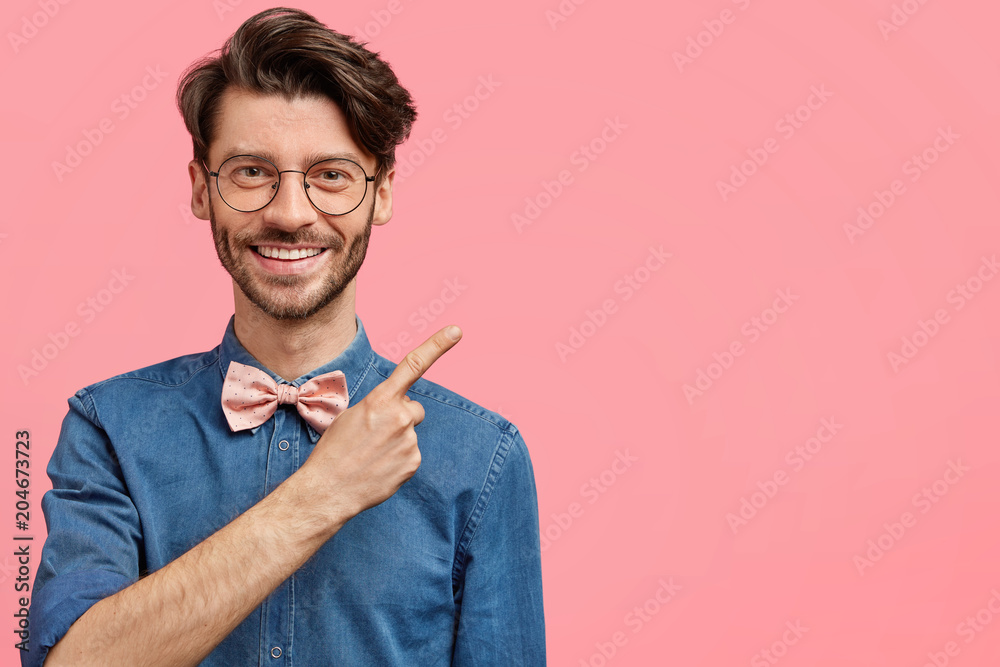 Pleasant looking unshaven young male with nice haircut, wears elegant clothes, indicates at blank pink space, shows place for your advertisement or promotional text. Positive hipster indoor. Pleasant looking unshaven young male with nice haircut, wears elegant clothes, indicates at blank pink space, shows place for your advertisement or promotional text. Positive hipster indoor.