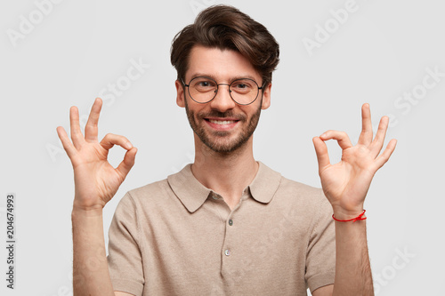 Cheerful delighted male student makes ok sign, being satisfied with results of passed exam, wears casual t shirt, smiles gently, isolated on white background. Glad unshaven happy man gestures