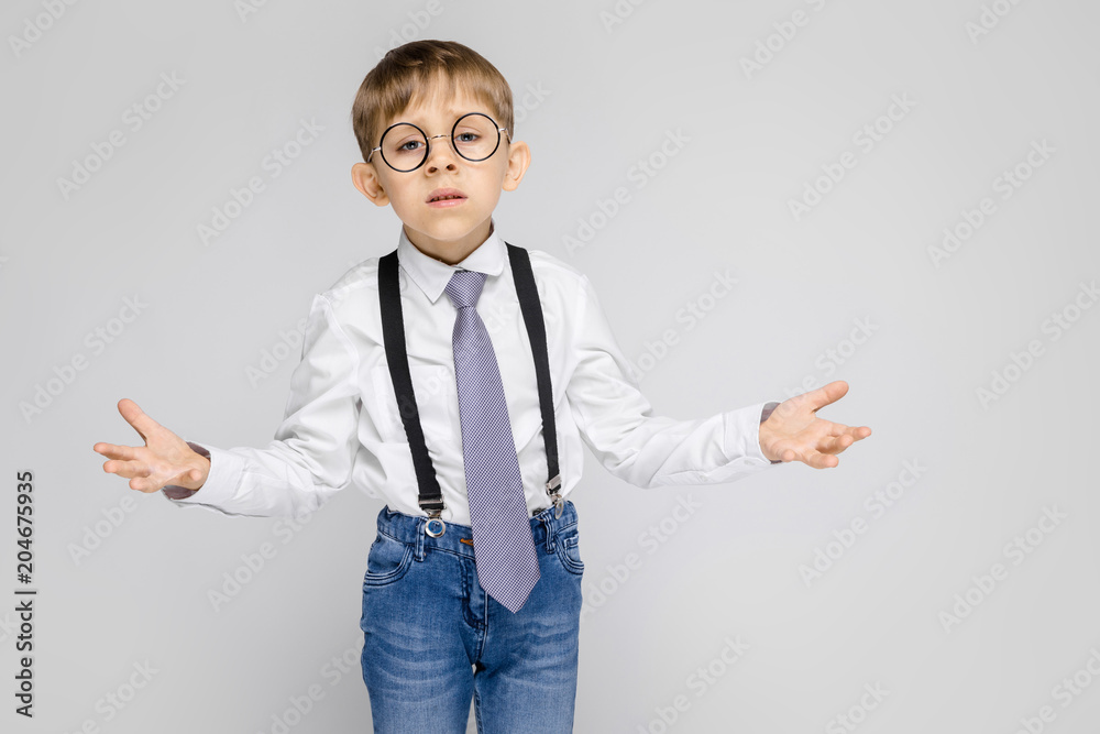 A charming boy in a white shirt, suspenders, a tie and light jeans stands on a gray background. The boy spread his hands in both directions A charming boy in a white shirt, suspenders, a tie and light jeans stands on a gray background. The boy spread his hands in both directions