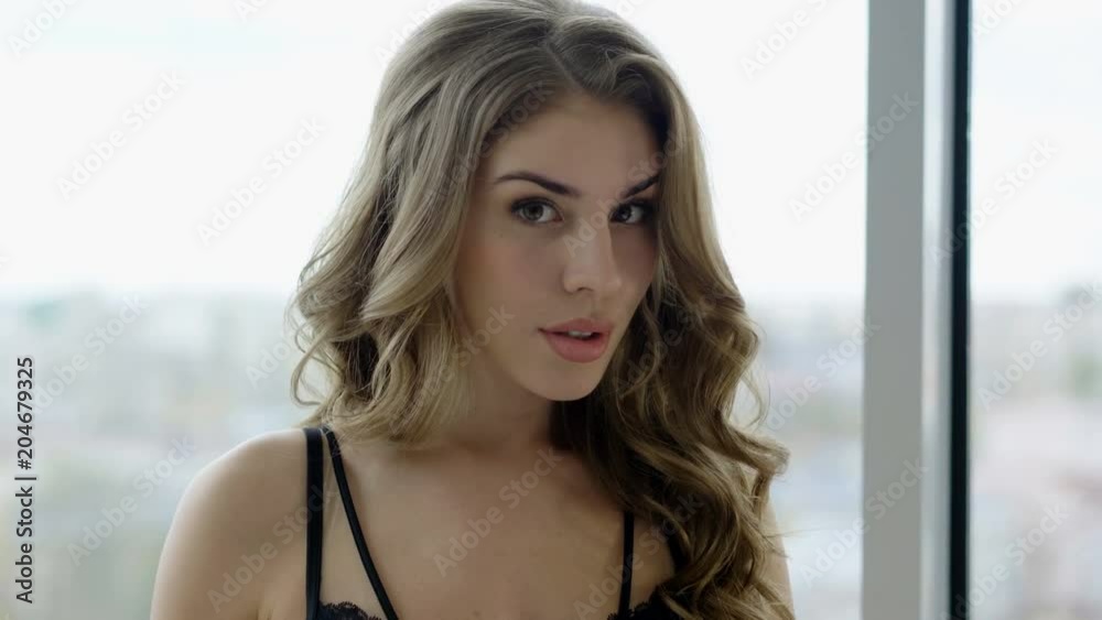 Portrait of beautiful blonde young woman in black lingerie standing at window in a luxury room. 4K