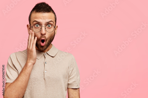 Horizontal shot of amazed attractive young bearded male looks with surprised expression, keeps mouth wide opened, poses against pink wall with copy space for your advertisement. Omg concept.