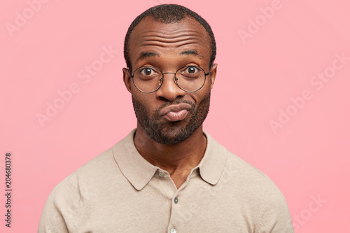 Headshot of handsome young man with hesitant look, looks in bewilderment, or puzzlement hears some doubtful information, poses on pink background. African American male being confused or uncertain