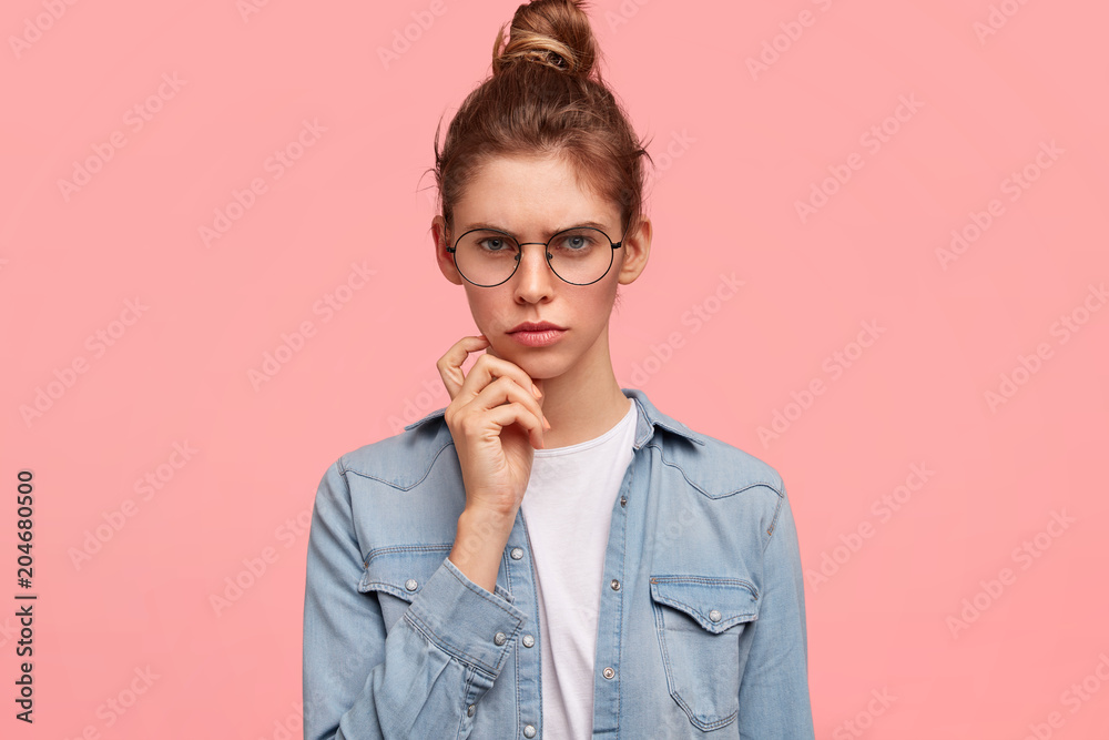 Unhappy young woman wears big spectacles, frowns face and looks suspiciously at camera, being displeased with negative news, isolated over pink background. People, attitude, emotions concept Unhappy young woman wears big spectacles, frowns face and looks suspiciously at camera, being displeased with negative news, isolated over pink background. People, attitude, emotions concept
