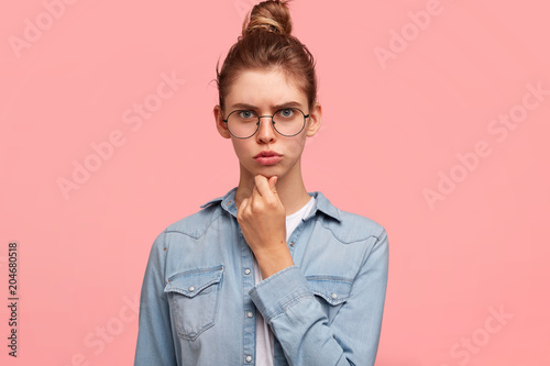 Photo of serious grumpy female model holds chin, looks seriously at camera, wears spectacles, denim jacket, has hair knot, expresses negative emotions. Displeased young woman writer, isolated on pink