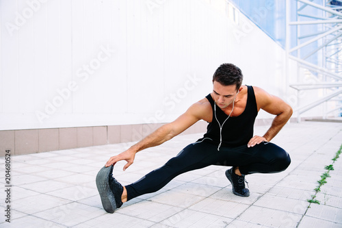 Attractive sportsman doing stretching exercises for legs, listening to music in headphones while working out outdoors. Wearing black sportswear.