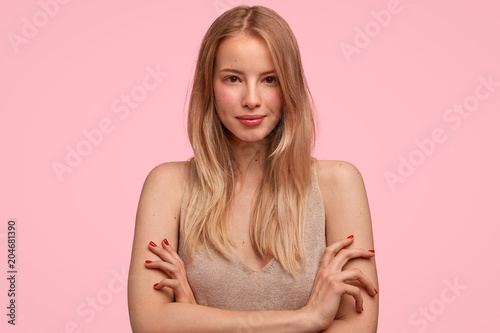 Confident beautiful Caucasian female with long hair, keeps arms folded, looks pleasantly and seriously at camera, listens something attentively, isolated on pink background. Self assured student
