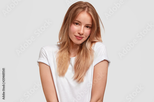 Horizontal shot of good looking Caucasian female model wears white t shirt in one tone with background, looks delightfully at camera, being satisfied with new purchase. Monochrome and trend.