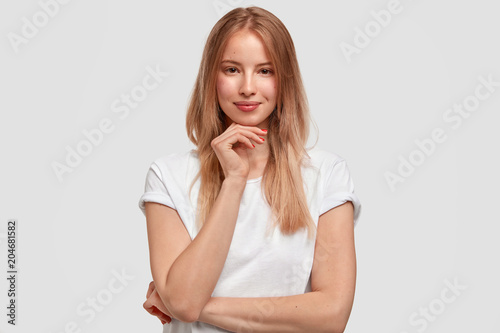Delighted female graduating student holds chin and looks happily directly into camera, wears casual white t shirt, isolated over studio background. Cute freelancer thinks about creating new project