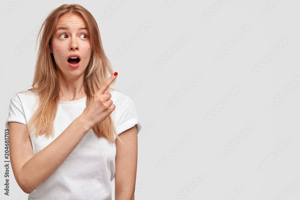 Lovely surprised female with appealing look, has long hair and shocked expression, indicates aside at blank copy space, wears casual white t shirt. Human facial expressions and advertisement concept Lovely surprised female with appealing look, has long hair and shocked expression, indicates aside at blank copy space, wears casual white t shirt. Human facial expressions and advertisement concept