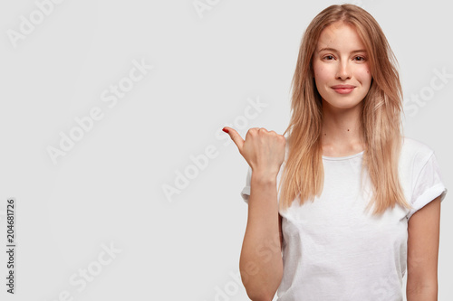 Photo of attractive delighted cute young European female indicates at blank white copy space for your advertisement or promotional text. Adorable woman seller advertises new product for buying.