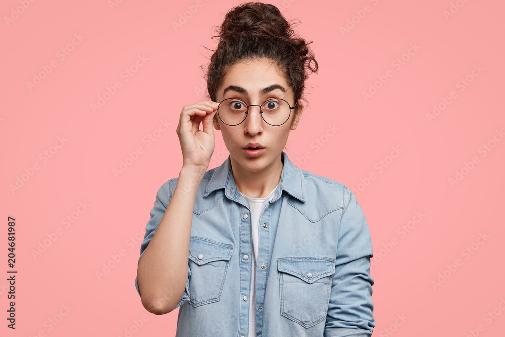 Surprised good looking young female with shocked expression, wears round glasses and denim shirt, looks with bated breath at camera, notices something unexpected, isolated over pink background Surprised good looking young female with shocked expression, wears round glasses and denim shirt, looks with bated breath at camera, notices something unexpected, isolated over pink background