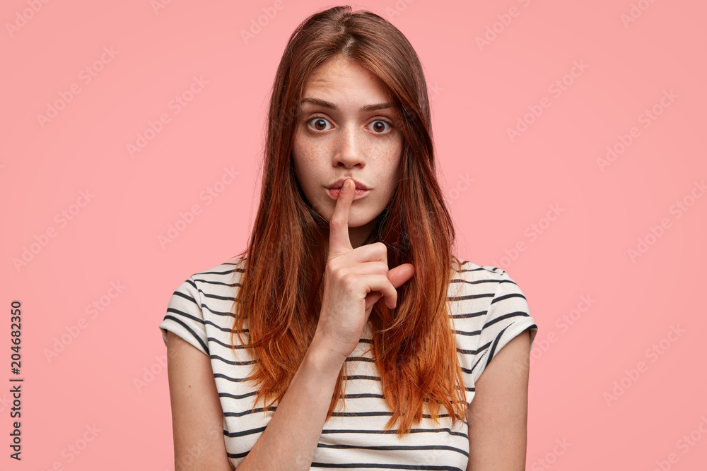 Top secret. Headshot of attractive freckled woman keeps fore finger on lips, asks to keep silence about her big secret, isolated on pink studio background. Pretty young female shows hush sign Top secret. Headshot of attractive freckled woman keeps fore finger on lips, asks to keep silence about her big secret, isolated on pink studio background. Pretty young female shows hush sign