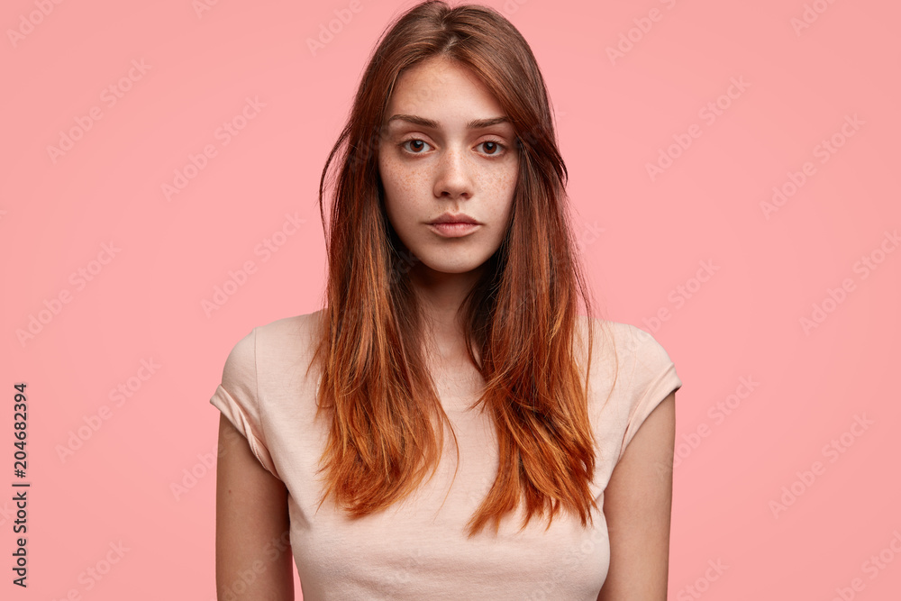 Candid shot of serious Caucasian female with freckled skin, dark eyes and long hair, wears casual clothing, poses against pink background. Adorable teenage girl poses in studio, being confident Candid shot of serious Caucasian female with freckled skin, dark eyes and long hair, wears casual clothing, poses against pink background. Adorable teenage girl poses in studio, being confident