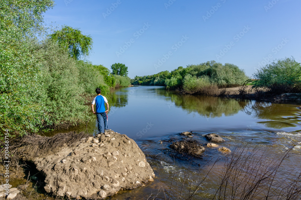 The teenager stands on top of a large stone boulder on the bank of the Sorraia River and looks at the river below. The river Sorraia in the summer - bright green vegetation,  blue sky