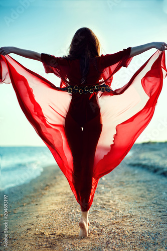 Beautiful woman with perfect body in trendy red chiffon beach cover up walking on tiptoe along the sea line towards the sunset holding the skirt of her gown flying in the wind. Beachwear concept photo