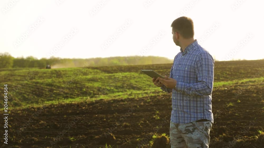Portrait of a beautiful young man farmer student working in the field with a tractor working in a tablet, happy, dirt in a shirt, the tractor plows a soil ground field. Concept ecology. Smart farming Portrait of a beautiful young man farmer student working in the field with a tractor working in a tablet, happy, dirt in a shirt, the tractor plows a soil ground field. Concept ecology. Smart farming