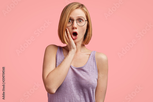 Stunned beautiful female journalist recieves unexpected news from colleague, wears casual purple t shirt and spectacles, poses against pink background. Stupefied young Caucasian woman in stupor