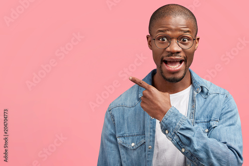 Handsome amazed African American bald man with bristle, wears fashionable denim shirt, indicates at blank copy space on pink background, advertises something. People, advertisement, emotions