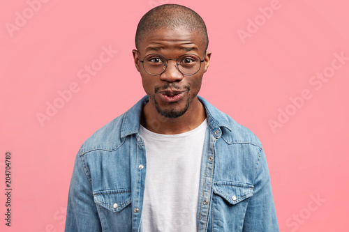 Bald funny middle aged African American male with curious glad expression, looks positively at camera, wears round glasses, stands against pink studo wall. People, ethnicity, emotions concept photo