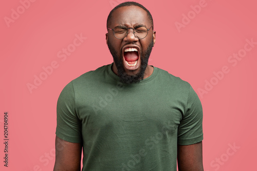 Crazy dark skinned male screams loudly in despair, keeps mouth wide opened and eyes closed, expresses negativeness, being furious and outraged, isolated over pink background. Anger, annoyance concept