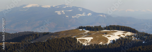 Breathtaking view of magnificent Carpathian mountain valley with dry grass and spots of snow on background of blurred mighty distant mountain range in early spring. photo