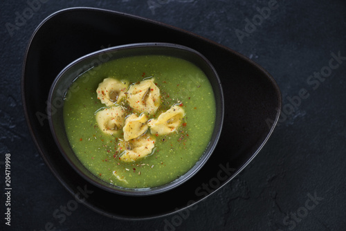 Black bowl with broccoli cream-soup and tortellini, top view on a black stone background, horizontal shot