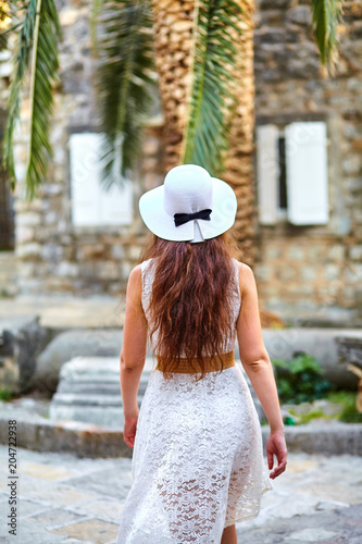 Fototapeta Naklejka Na Ścianę i Meble -  Young brown-haired woman in white hat and dress walking the streets of the old city